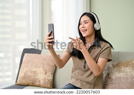 Joyful and beautiful young Asian female in casual clothes wearing headphones, enjoys talking with her friends on video call through her smartphone while relaxing in her living room.