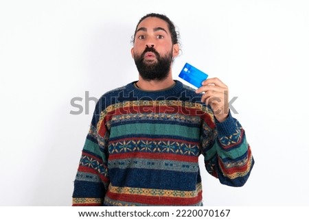 Photo of young hispanic bearded man wearing knitted sweater over white background amazed shocked hold credit card payment