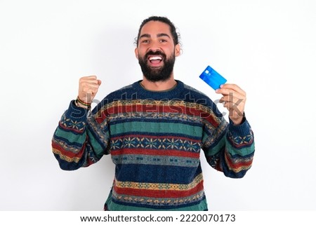 Photo of lucky impressed young hispanic bearded man wearing knitted sweater over white background arm fist holding credit card. Celebrated