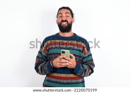 Photo of astonished crazy young hispanic bearded man wearing knitted sweater over white background hold smartphone dislike feedback concept