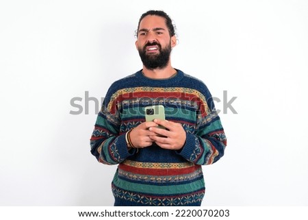 Photo of astonished crazy young hispanic bearded man wearing knitted sweater over white background hold smartphone dislike feedback concept
