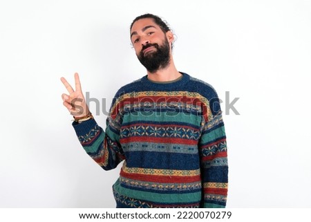 young hispanic bearded man wearing knitted sweater over white background makes peace gesture keeps lips folded shows v sign. Body language concept