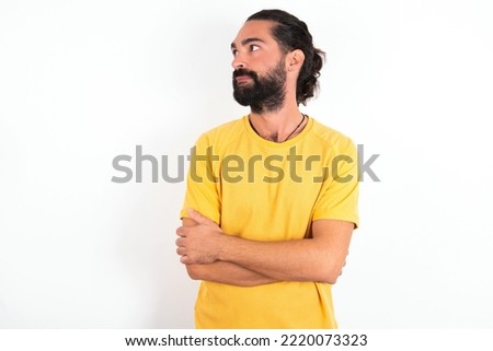 Pleased young hispanic bearded man wearing yellow T-shirt over white background keeps hands crossed over chest looks happily aside