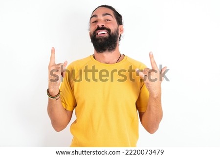young hispanic bearded man wearing yellow T-shirt over white background makes rock n roll sign looks self confident and cheerful enjoys cool music at party. Body language concept.