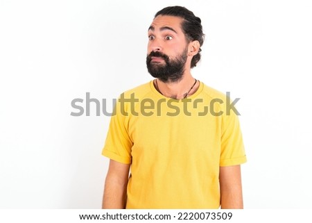 young hispanic bearded man wearing yellow T-shirt over white background stares aside with wondered expression has speechless expression. Embarrassed model looks in surprise