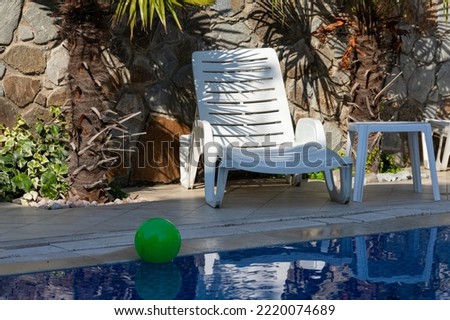 White sun lounger by the pool with blue water and a green ball, against the backdrop of palm trees.