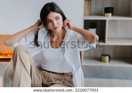 Upset Korean girl in casual sitting in chair  at home over blurry fireplace and bookshelf, looking aside adjusts collar on shirt. Pretty Asian student woman overloaded, learning home. Luxury life.