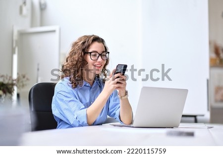 Young happy business woman, smiling beautiful professional lady worker looking at smartphone using cellphone mobile cellular tech working at home or in office checking cell phone sitting at desk.