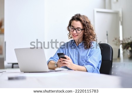 Young happy business woman, smiling pretty professional businesswoman worker looking at smartphone using cellphone mobile technology working at home or in office checking cell phone sitting at desk. Royalty-Free Stock Photo #2220115783