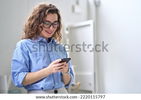 Smiling happy young business woman manager holding smartphone working standing in office using mobile cell phone, checking financial banking apps, browsing online on cellphone typing on cellular. Royalty-Free Stock Photo #2220115797