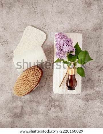 Spa treatment flat lay with bottle of organic oil essence collagen. Dropper bottle with natural serum on a white towel with lilac flowers, massage brush and loofahs on a grey textured background