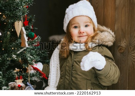 cute little girl in knitted white hat stand at porch of country house and decorate Christmas tree for celebration New Year and Christmas holidays for children outdoor