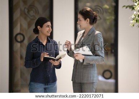 Businesswomen stand in office hallway and talking, boss gives task to Indian secretary at workday in modern workspace. Personal assistant or intern, listens to female chief. Mentoring, apprenticeship Royalty-Free Stock Photo #2220172071