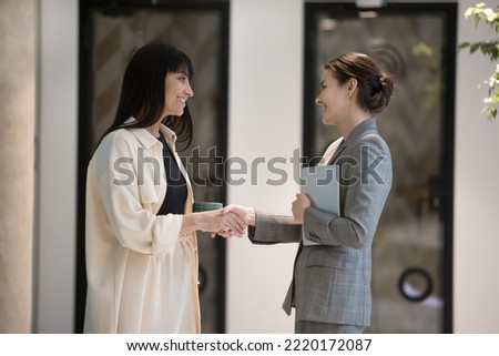 Two attractive business women, colleagues greeting, handshaking while talking standing in office hall. Successful businesswomen, professionals shaking hands, express respect, meet in company hallway Royalty-Free Stock Photo #2220172087
