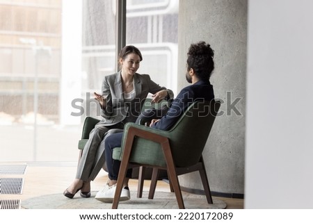 Two multinational workmates talking seated on armchairs in modern office, share opinion and thoughts, discuss common project, share solution looking interested in conversation. Business, communication Royalty-Free Stock Photo #2220172089