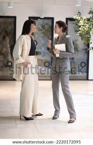 Two businesswomen chatting, meet together in hallway, share opinions and professional information, discuss project having business conversation or informal talk with tablet and coffee mug in office Royalty-Free Stock Photo #2220172095