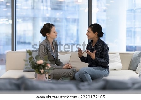 Multiethnic businesswomen, Caucasian and Indian female workmates sit on cozy sofa in modern office lobby lead informal conversation during break at workspace. Communication at workplace, discussion Royalty-Free Stock Photo #2220172105