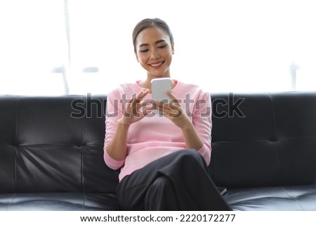 Happy Asian woman sitting on the sofa playing mobile phone in the living room at home.