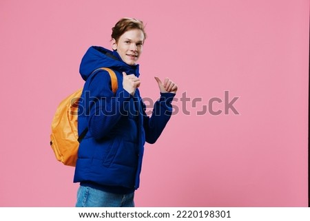 funny funny red-haired guy shows standing sideways to the camera with his finger on a bright backpack on his back while standing in a warm winter jacket on a light blue background