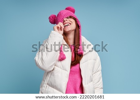 Playful woman in winter coat pulling her funky hat on yes while standing against blue background