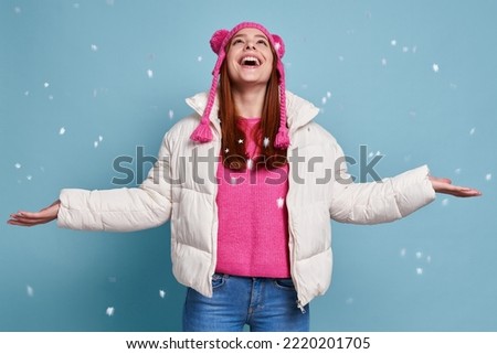 Happy woman in winter coat keeping arms outstretched while snow falling against blue background