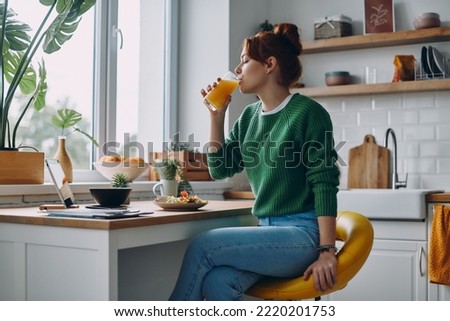 Beautiful redhead woman drinking juice while having lunch at the domestic kitchen Royalty-Free Stock Photo #2220201753