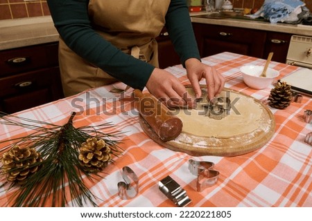 Close-up female confectioner in beige chef's apron, putting cookies cutters on a rolled out gingerbread dough, preparing delicious homemade pastries for Christmas holidays in the kitchen at home