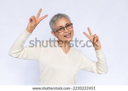 Portrait of cheerful senior woman making peace gesture. Mature Caucasian woman wearing eyeglasses and white jumper showing victory sign and smiling. Fun and happiness concept