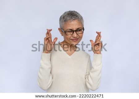 Portrait of deceitful senior woman making liar gesture. Mature Caucasian woman wearing eyeglasses and white jumper crossing fingers in hope over white background. Liar, hope, wish concept