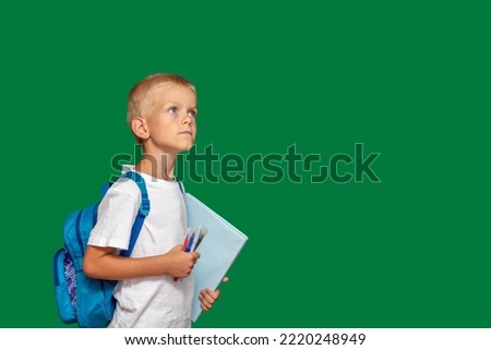 Boy with backpack, book and markers in his hands, looks up. Green background with space for text. Selective focus. Picture for articles about children, school, education.