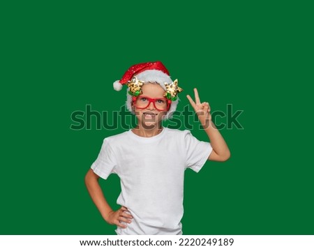 Boy in Santa Claus hat and glasses, shows peace sign with his hand and smiles. Green background with space for text. Selective focus. Picture about children, holidays, Christmas.