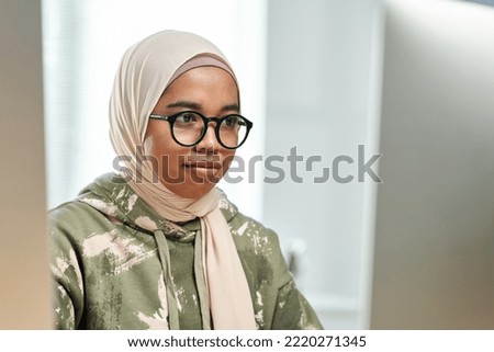 Young Muslim black woman in hijab and eyeglasses sitting in front of computer screen in office while developing new software