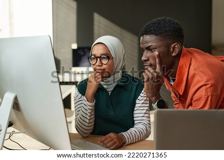 Two young serious intercultural IT engineers looking at data on computer screen or waiting for web page uploading at meeting in office