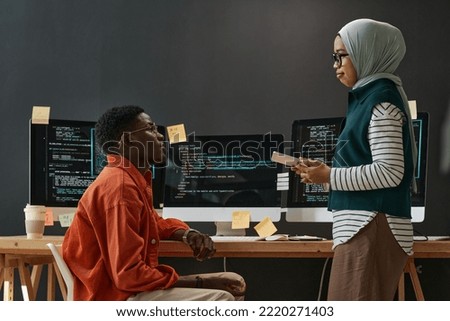 Two young confident multicultural interns in casualwear looking at each other during discussion of working points at meeting