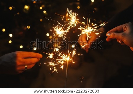 Happy New Year! Friends celebrating with burning sparklers in hands against christmas tree lights in dark room. Hands holding fireworks on background of stylish decorated illuminated tree. Moody Royalty-Free Stock Photo #2220272033