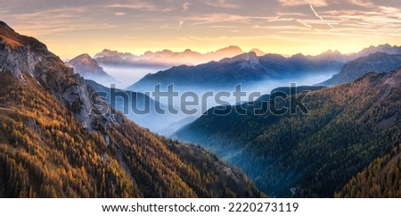 Mountains in low clouds at sunset in autumn in Dolomites, Italy. Landscape with alpine mountain hills in fog, orange trees and grass in fall, colorful sky with golden sunbeams. Aerial view. Panorama Royalty-Free Stock Photo #2220273119