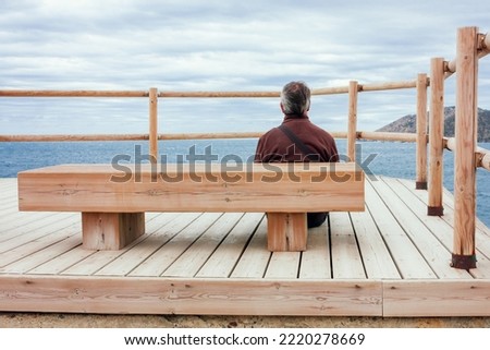 A man is sitting in front of a bench contemplating the sea. He is on his back. Sitting on the floor of a wooden balcony.