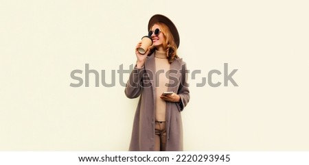 Portrait of stylish beautiful young woman model with cup of coffee wearing brown round hat and coat on beige background