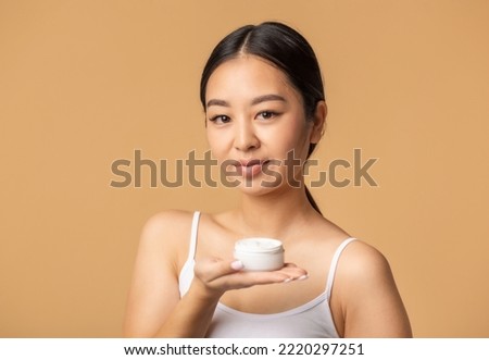 Beautiful korean woman holds moisturizer face cream standing on a beige background, looking at the came