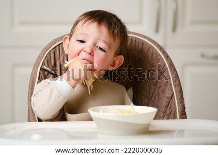 A funny child is eating a grated apple with his mouth full while sitting on a kitchen chair. Hungry baby boy shoves food in his mouth, humor. Kid aged one year and three months