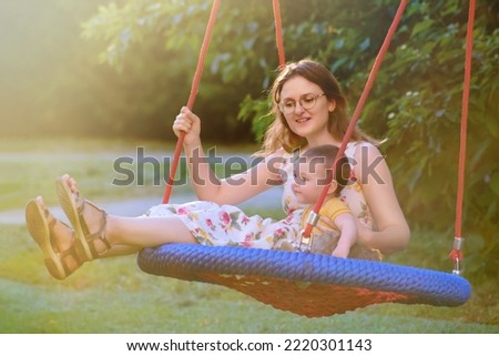 A mother woman together with a toddler baby swings on a swing in the park. Mom and child are sitting on a swing in nature