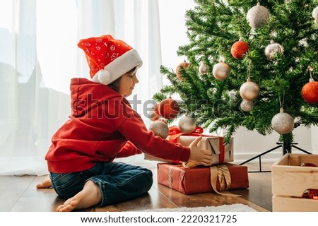 Charming boy in a santa hat takes out Christmas gifts from under the New Year tree and laughs