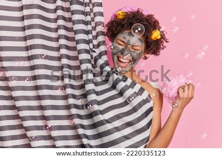 Hygiene procedures and routines concept. Joyful curly haired woman enjoys showering applies beauty facial mask for skin care exclaims from joy poses behind striped shower curtain against pink wall Royalty-Free Stock Photo #2220335123
