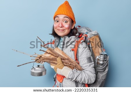 Horizontal shot of pretty teenager wears hat windbreaker and gloves has active rest holds kettle wood carries rucksack with necessary equipment isolated over blue background. People recreation concept
