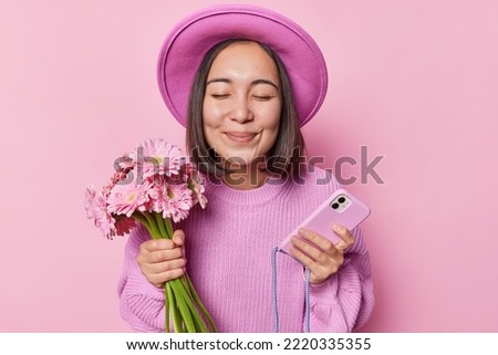 Horizontal shot of pretty dark haired Asian woman wears hat and knitted jumper holds bouquet of flowers modern smartphone goes to friends birthday keeps eyes closed isolated over pink background
