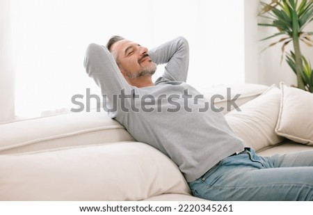 Relaxed man enjoying rest lying on comfortable couch with the hands on the head in living room at home. Calm middle-age man breathing fresh air enjoying no stress free peaceful weekend in house. Royalty-Free Stock Photo #2220345261