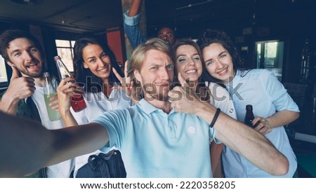 Group of happy colleagues are taking selfie together, young man is holding camera and posing, his coworkers are holding drinks in bottles, laughing and looking at camera.