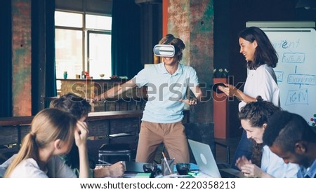 Multi-ethnic group of young people is having fun with augmented reality glasses, guy is using them moving hands and body, laughing woman is working with tablet. Royalty-Free Stock Photo #2220358213