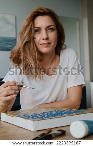 Closeup portrait of happy creative young woman smiling and looking directly to the camera during painting on canvas in her studio. Person holding paintbrush in workshop Royalty-Free Stock Photo #2220395187