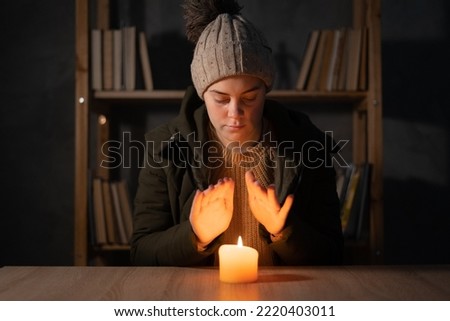 Freezing young woman in winter clothes warms her hands on lights with candles. suffers in no heating and no electricity during an energy crisis in Europe causing blackouts. copy space.